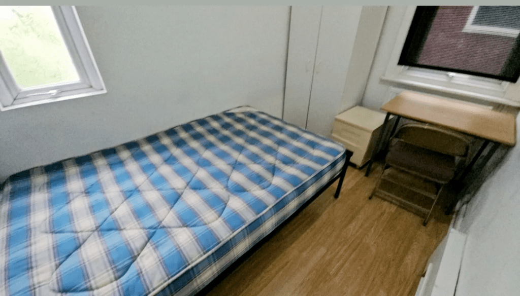 A stark room with an uncovered mattress, a desk, a tiny chest of drawers, and a looming wardrobe. All are made of MDF, and none of them are the same colour. The vibe is 'prison cell'.